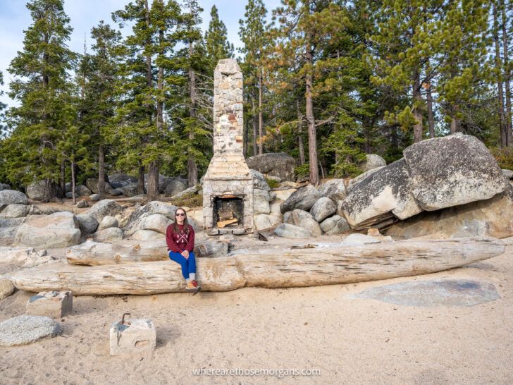 Hiker in maroon sweater sat on a log in front of a stone chimney on Chimney Beach in Lake Tahoe in late afternoon