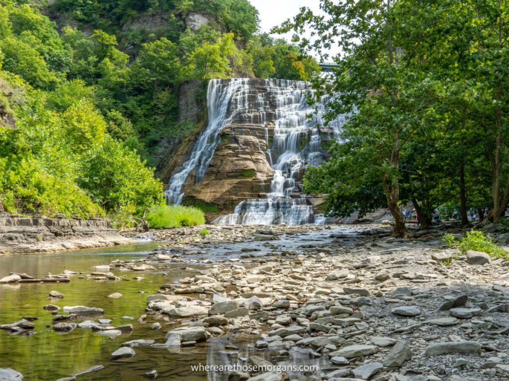 Popular waterfall flowing gently over a rocky cliff into a creek with low water and trees with lush green leaves at Ithaca Falls NY