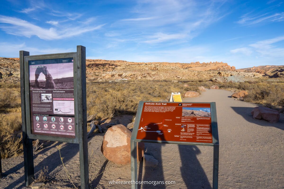 Trailhead for a hike with information signs in late afternoon leading to a dirt path cutting through rocks and vegetation