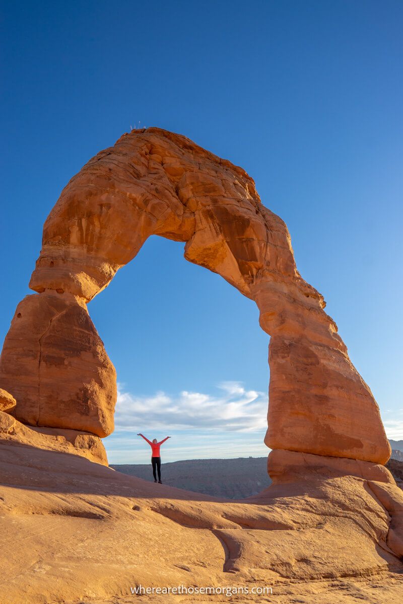 Hiker stood underneath Delicate Arch in Utah with arms raised for perspective