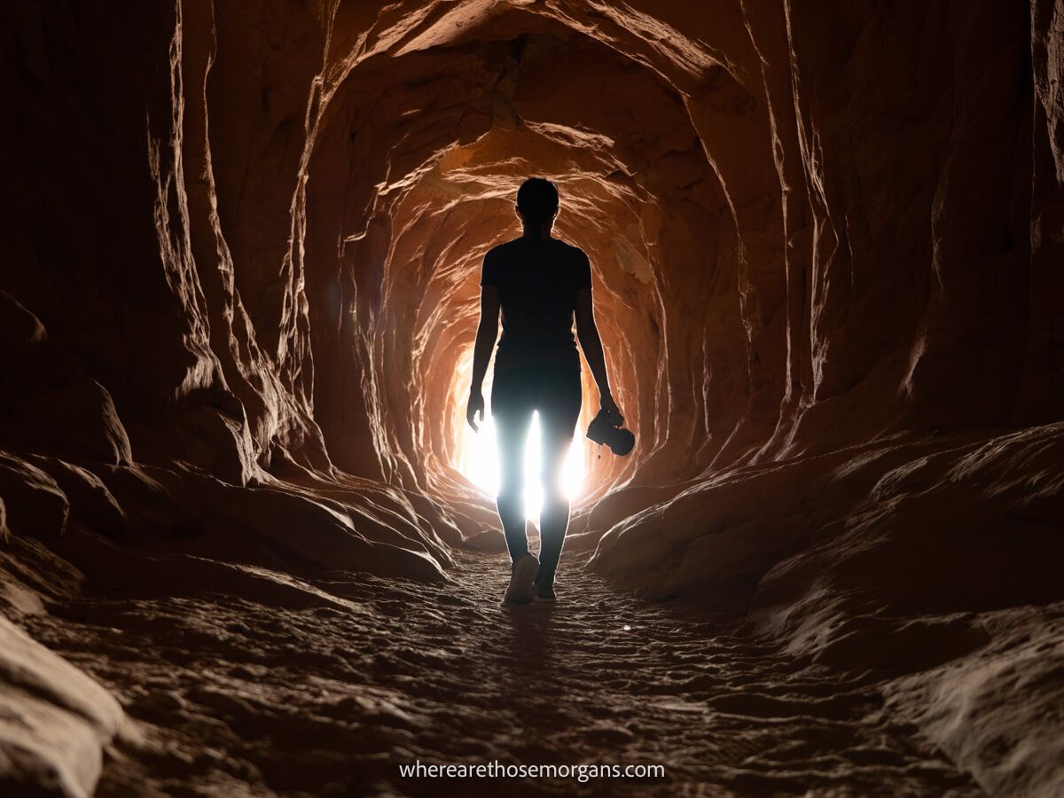 Hiker with camera walking through sandstone tunnel silhouetted by light