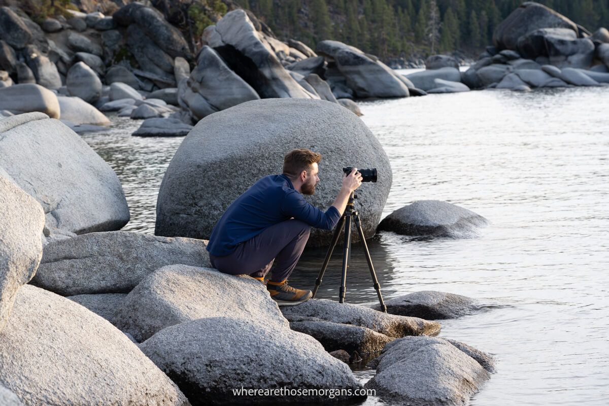 Photographer crouching with tripod and camera on a boulder next to water