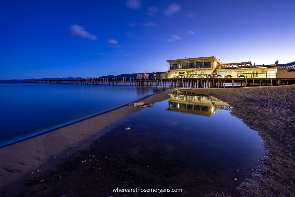 Pier and boathouse lit up at dawn reflecting in shallow pool of water on a beach