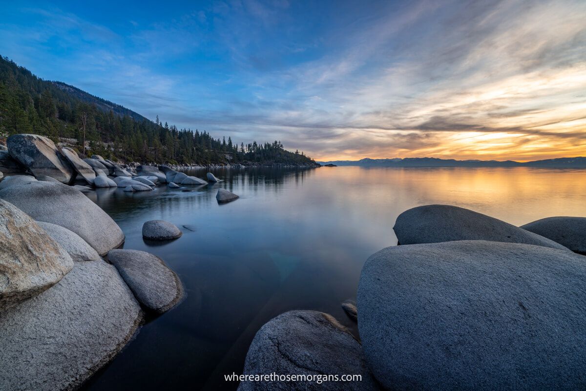 Large boulders in a smooth lake with trees on a hill to the left and a sunset with colorful clouds to the right