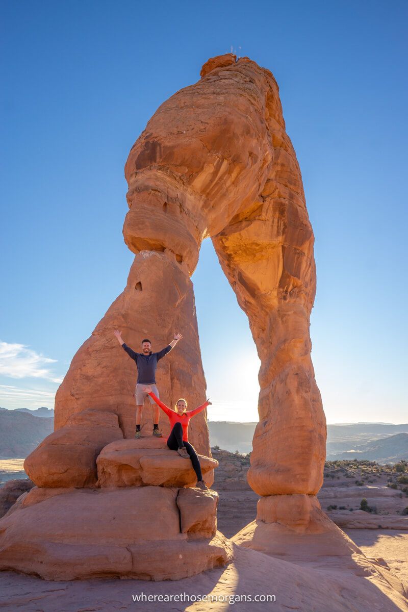 Two hikers sat and stood in front of Delicate Arch in Arches on a sunny day