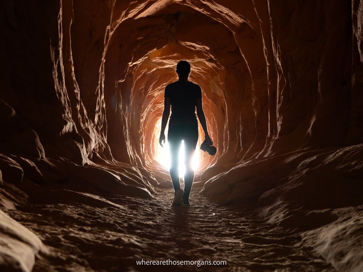 Hiker holding camera walking through sandstone tunnel silhouetted against the light