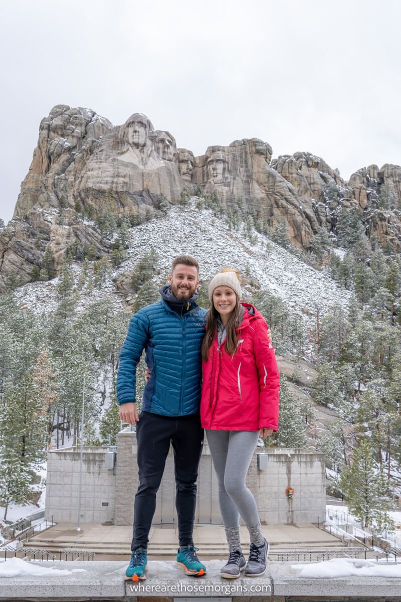 Couple standing in front of Mount Rushmore in winter clothes with snow on the ground