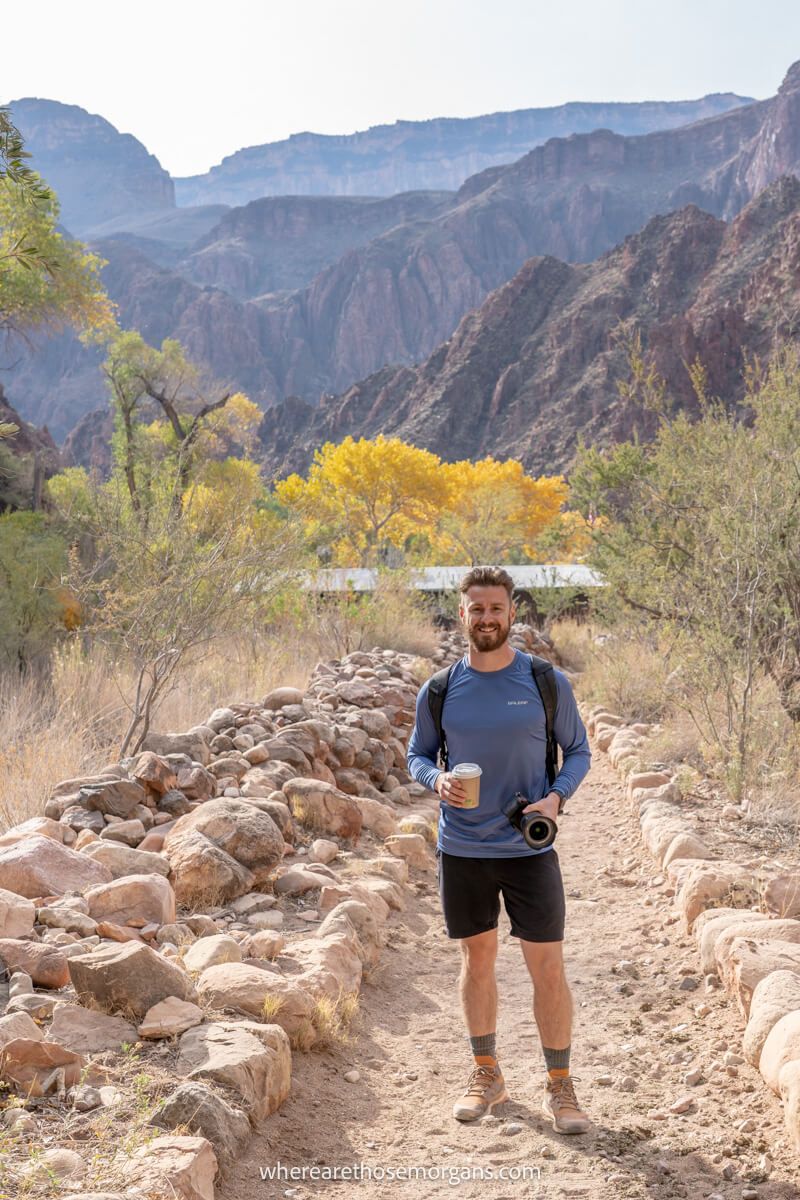 Hiker in shorts and long sleeved shirt holding a camera and hot chocolate at Phantom Ranch in the bottom of the Grand Canyon on a sunny day