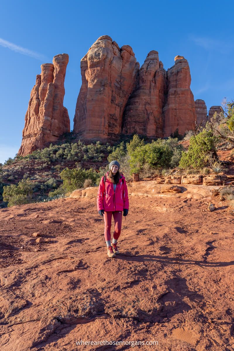 Tourist in winter coat walking on a red rock path with tall sandstone spires behind on a clear day