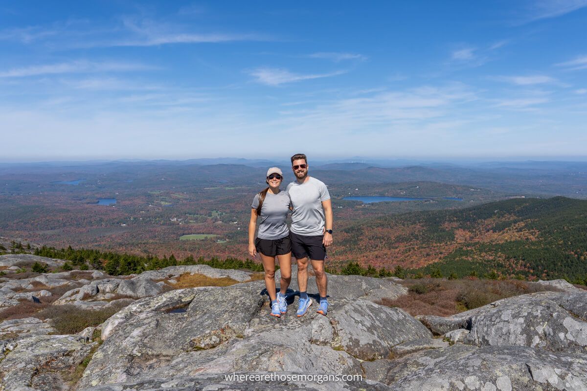 Couple standing together for a summit photo at the top of a mountain with far reaching views and blue sky behind