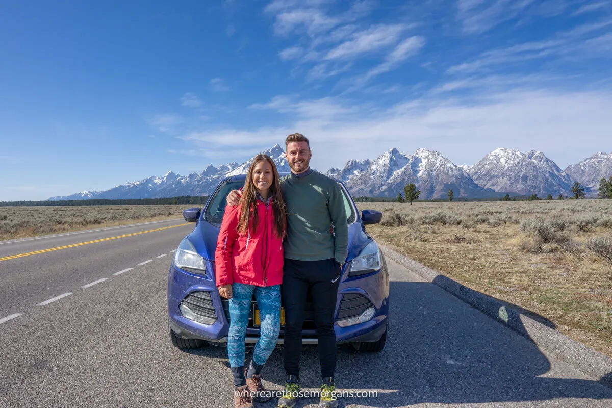 Couple standing against a blue car on a road with mountains in the background