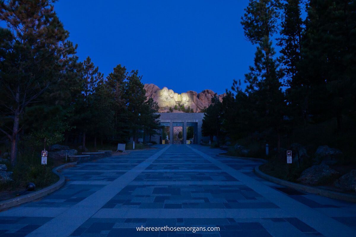 Photo showing Mount Rushmore lit up at night from a distance with the path and trees leading up to the entrance covered in dark shadows