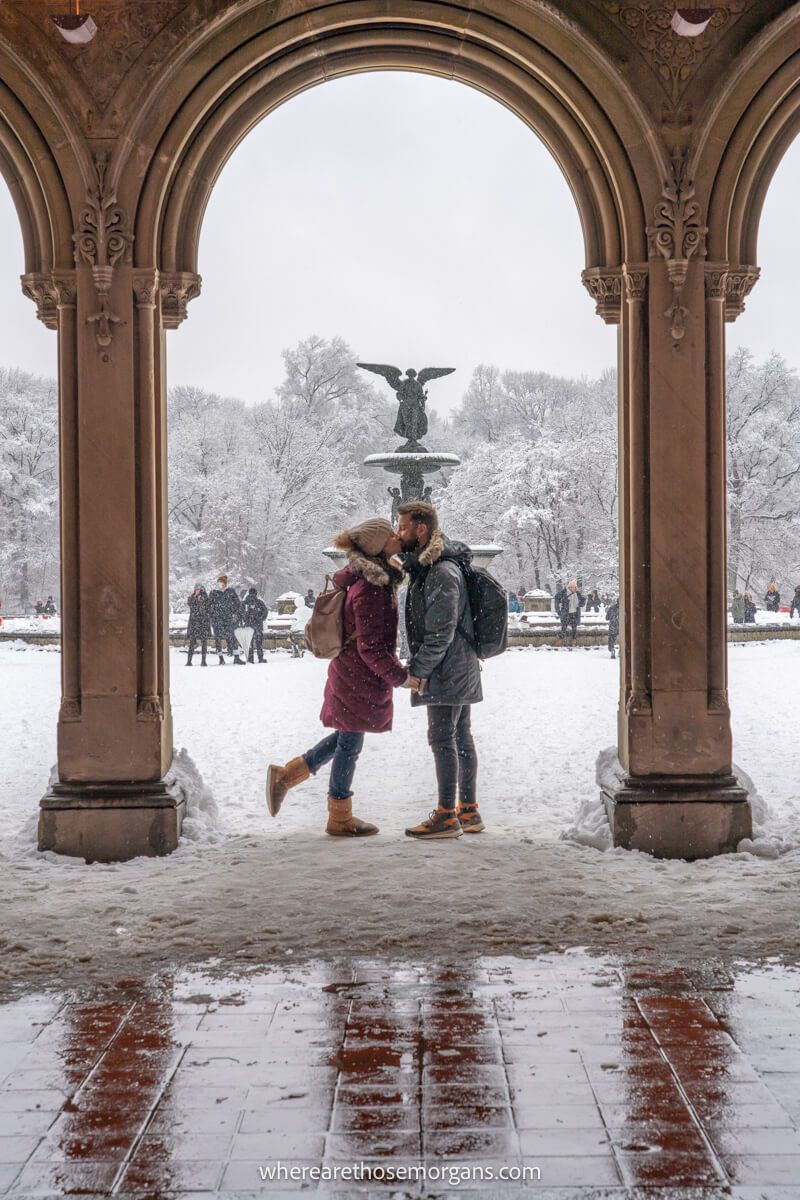 Couple in winter coats kissing on snow between columns in front of a fountain on a cloudy day