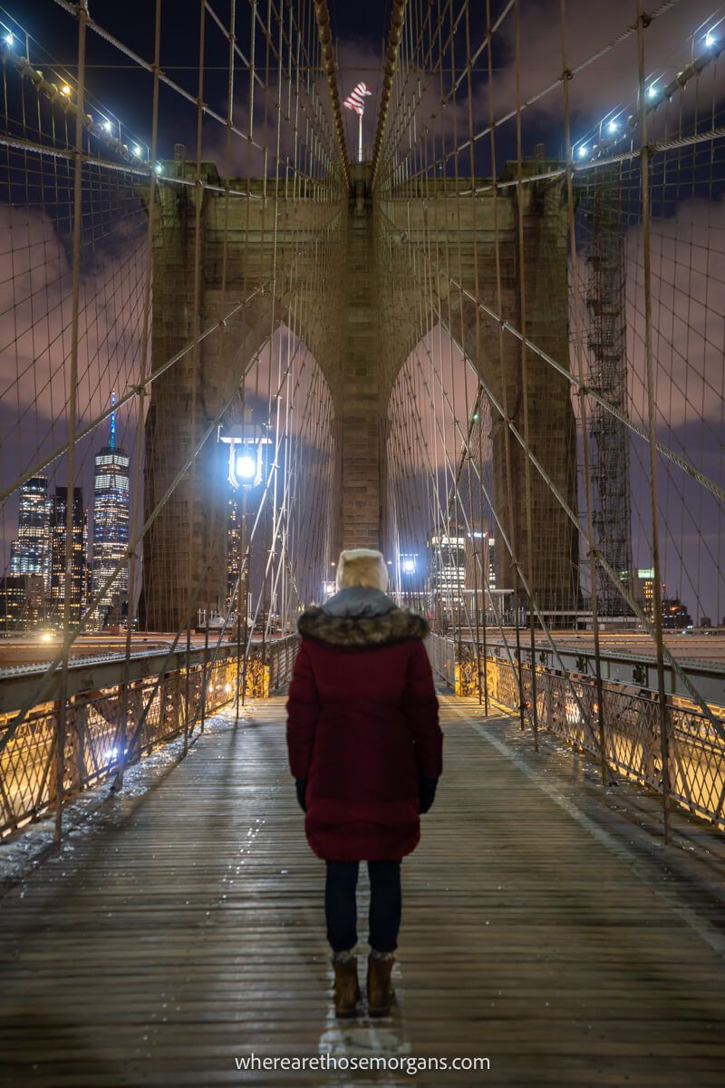 Tourist in winter coat and hat walking across the Brooklyn Bridge in NYC at night with nobody else on the walkway