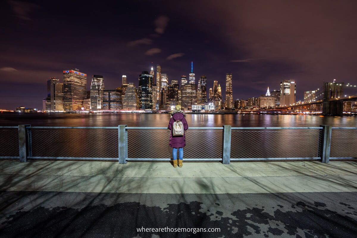 Tourist in winter coat standing against a fence looking at a river and city lit up at night