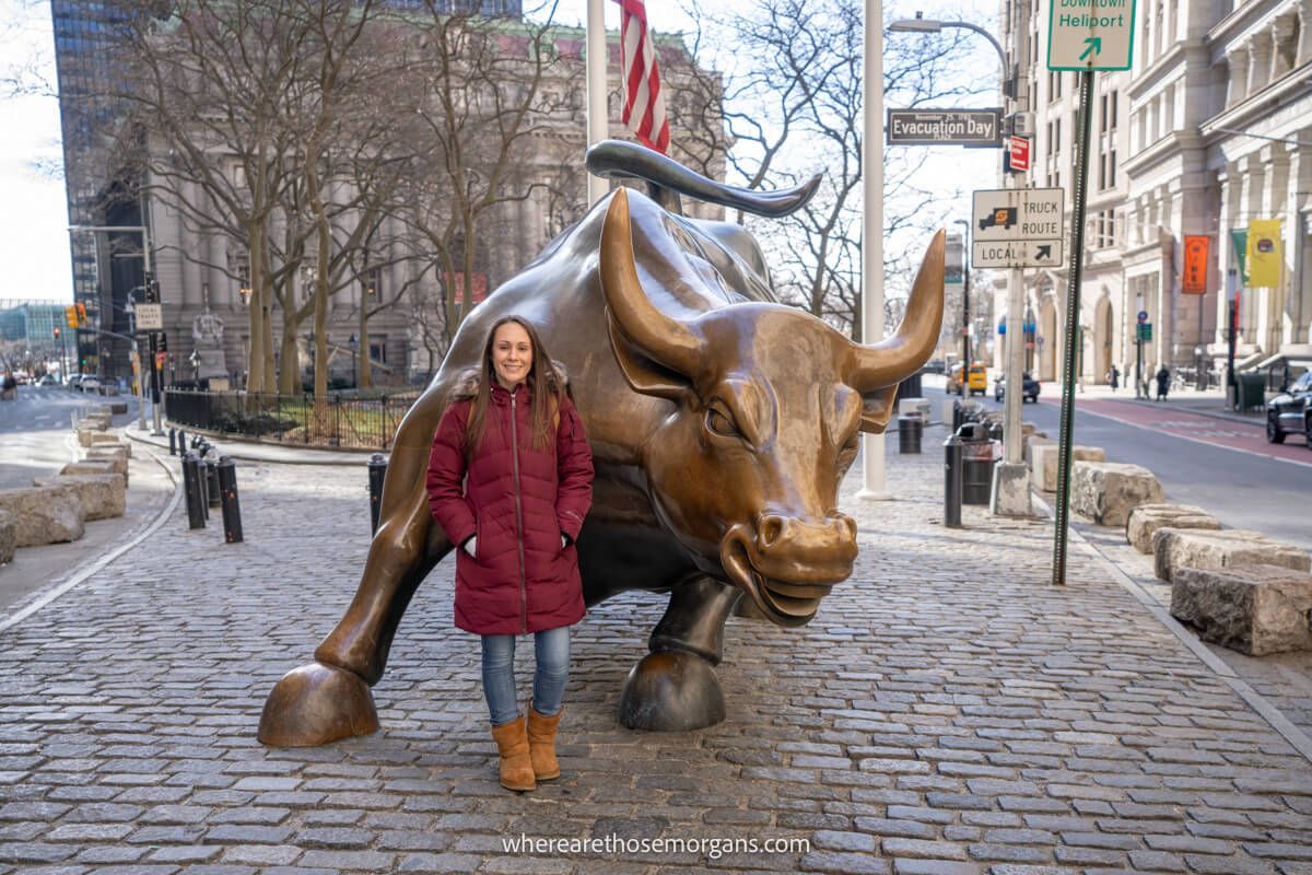 Photo of a tourist in winter coat smiling next to a large statue of a bull in NYC