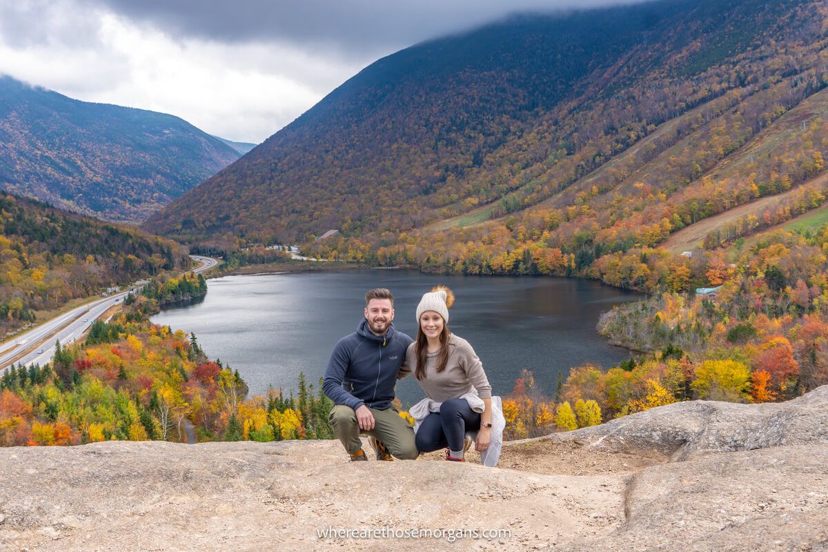 Hikers crouching next to each other on a rocky surface for a photo with a lake and mountains covered in colorful leaves behind