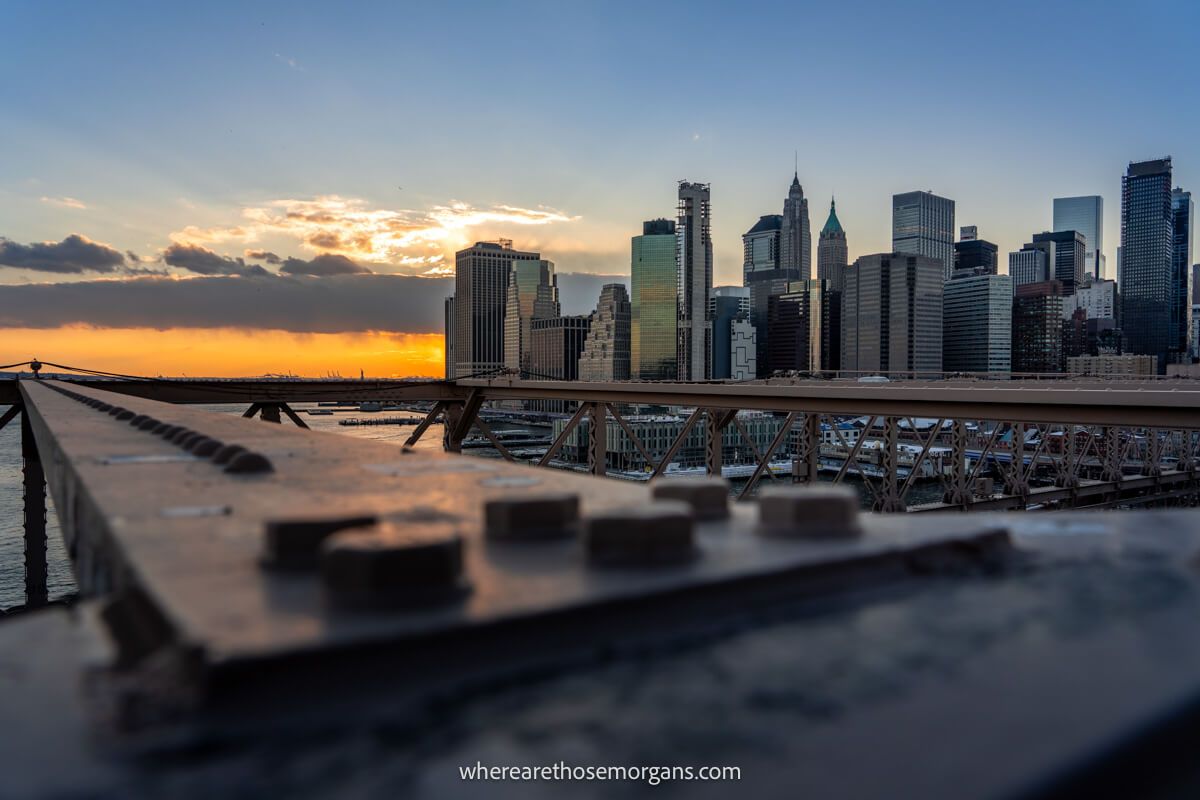 The Lower Manhattan skyline of skyscrapers at sunset from Brooklyn Bridge with a long metal girder in the foreground
