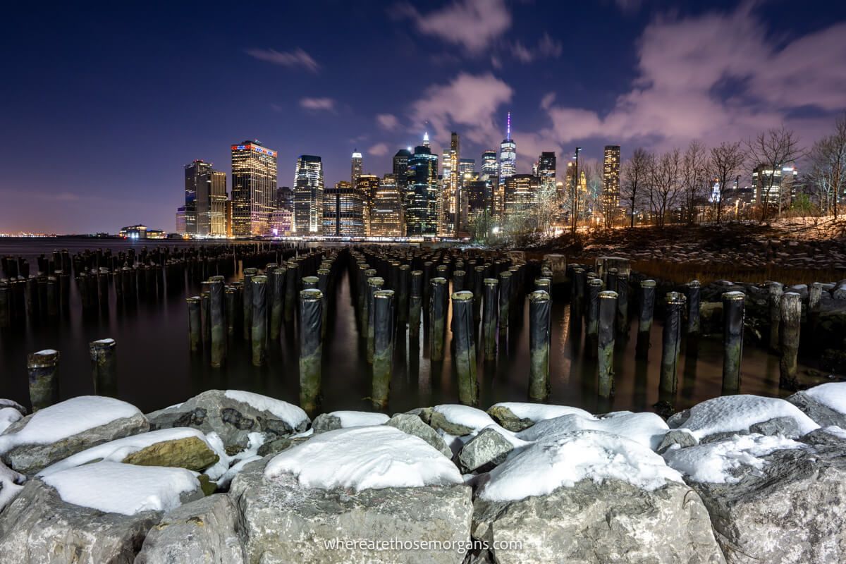 Snow covered stones in front of a harbor filled with wooden poles and city skyscrapers lit up at night