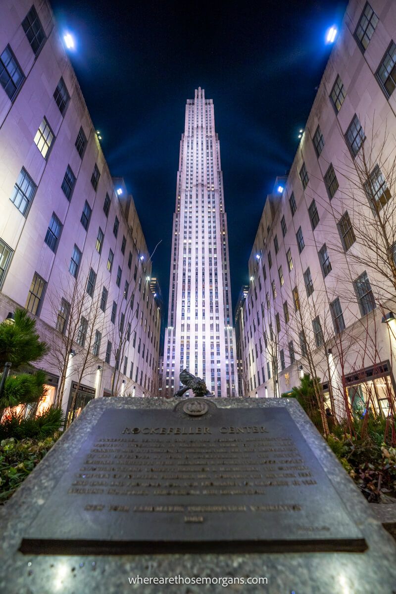 Tall narrow building bursting into the sky lit up purple and blue at night by lights on two flanking buildings with a plaque in the foreground
