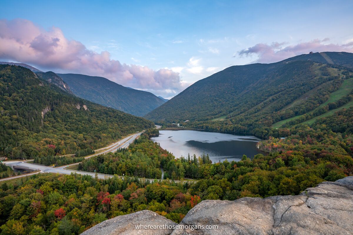 View over Echo Lake and rolling hills covered in trees with pink colored clouds from a rocky summit viewpoint called Artists Bluff in New Hampshire