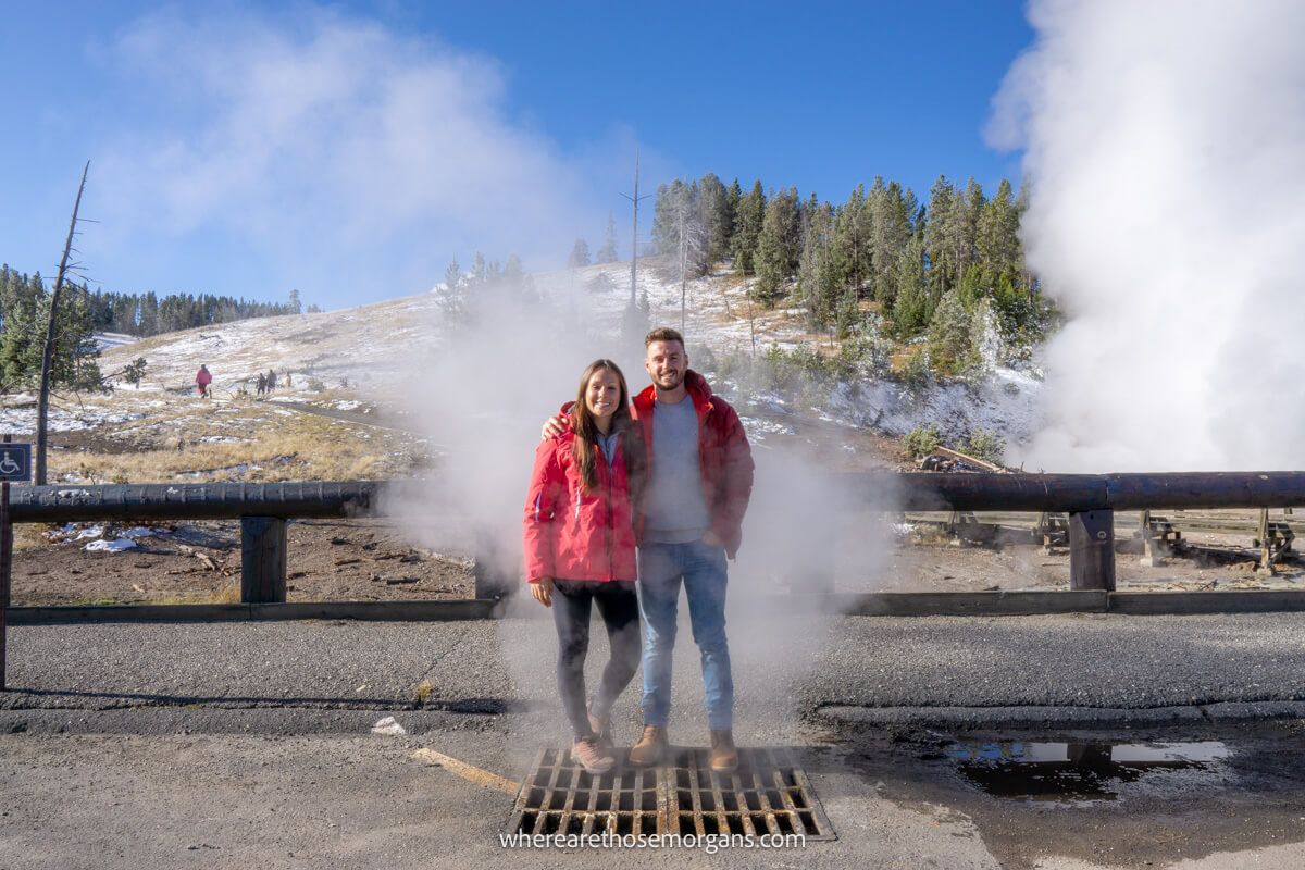 Couple standing together in winter clothes on top of a metal grate with steam coming up from below and more steam billowing out of geysers behind at Yellowstone National Park in October
