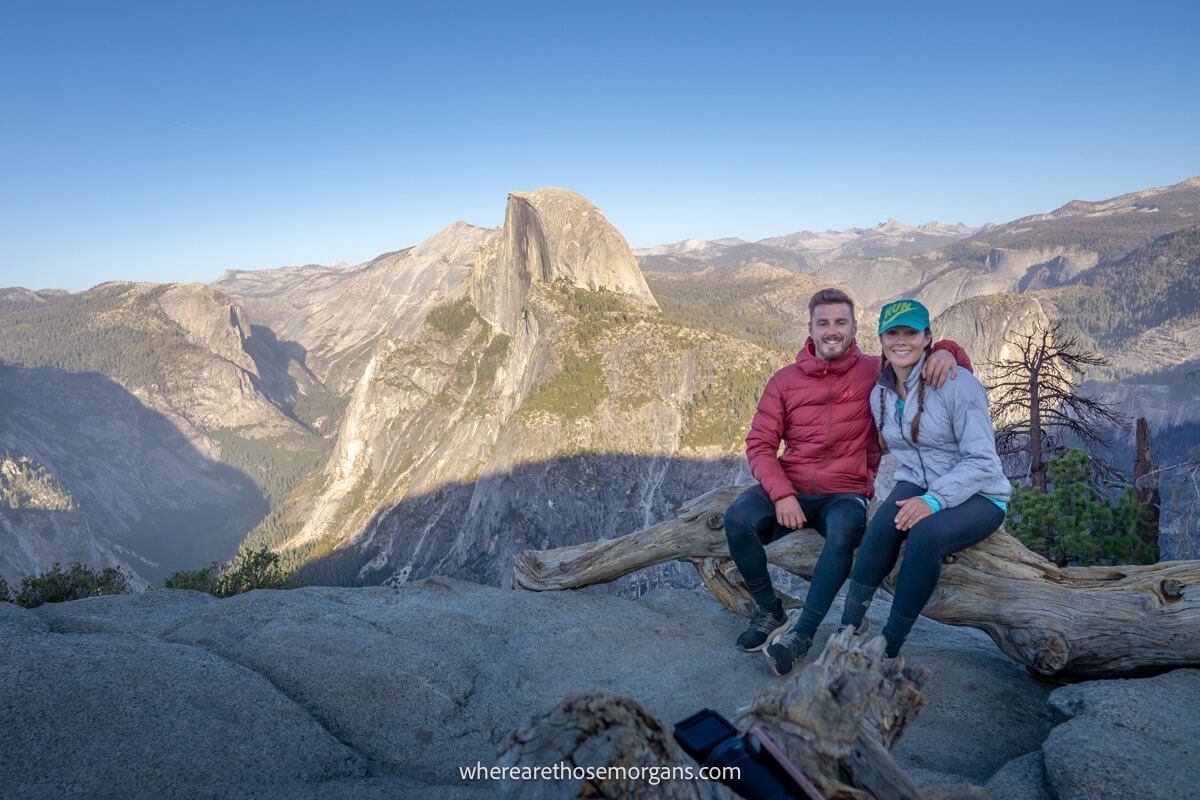 Couple sat together in winter coats on a log at a rocky summit with far reaching wide open views over Half Dome, granite peaks and Sierra Nevada mountains in California