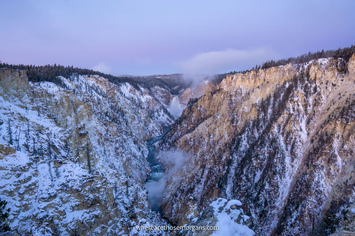 Photo of a deep V-shaped canyon leading to a powerful waterfall in the distance with snow on the slopes and pink colors in the sky at dawn