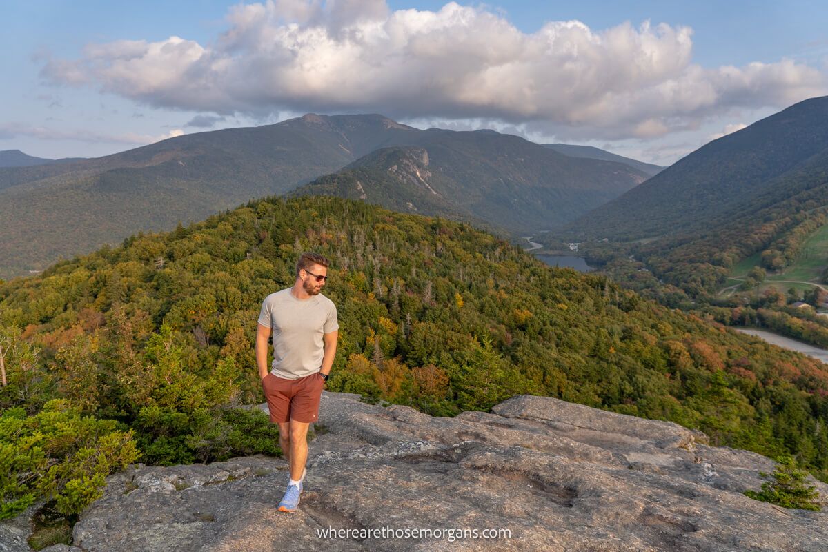 Hiker walking across a rocky summit with far reaching views over rolling hills covered in trees at sunset