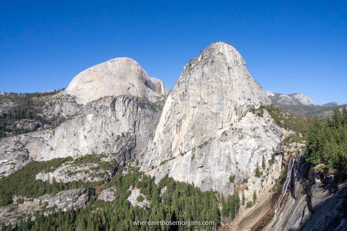 Landscape photo of a nearly dry waterfall next to tall granite domes with lots of evergreen trees on a clear day