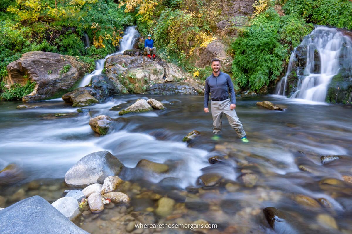 Small waterfalls on the far side of a river with a hiker sat on a rock and another hike stood in the river for a photo at Big Springs the end of the bottom up day hike in The Narrows
