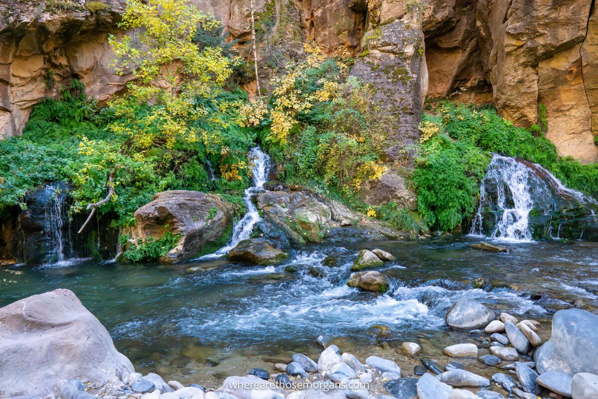 Three small waterfalls flowing into a river with small stones and a towering red rock cliff behind