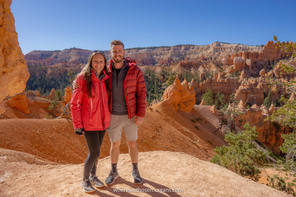 Two hikers stood together in winter coats for a photo on the Queens Garden Navajo Loop Trail in Bryce Canyon with far reaching views over sandstone cliffs and hoodoos