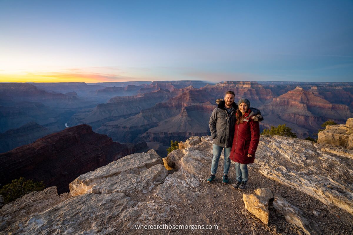 Couple standing together for a photo in heavy winter coats on a rocky cliff with distant views over Grand Canyon South Rim at sunset with colors in the sky