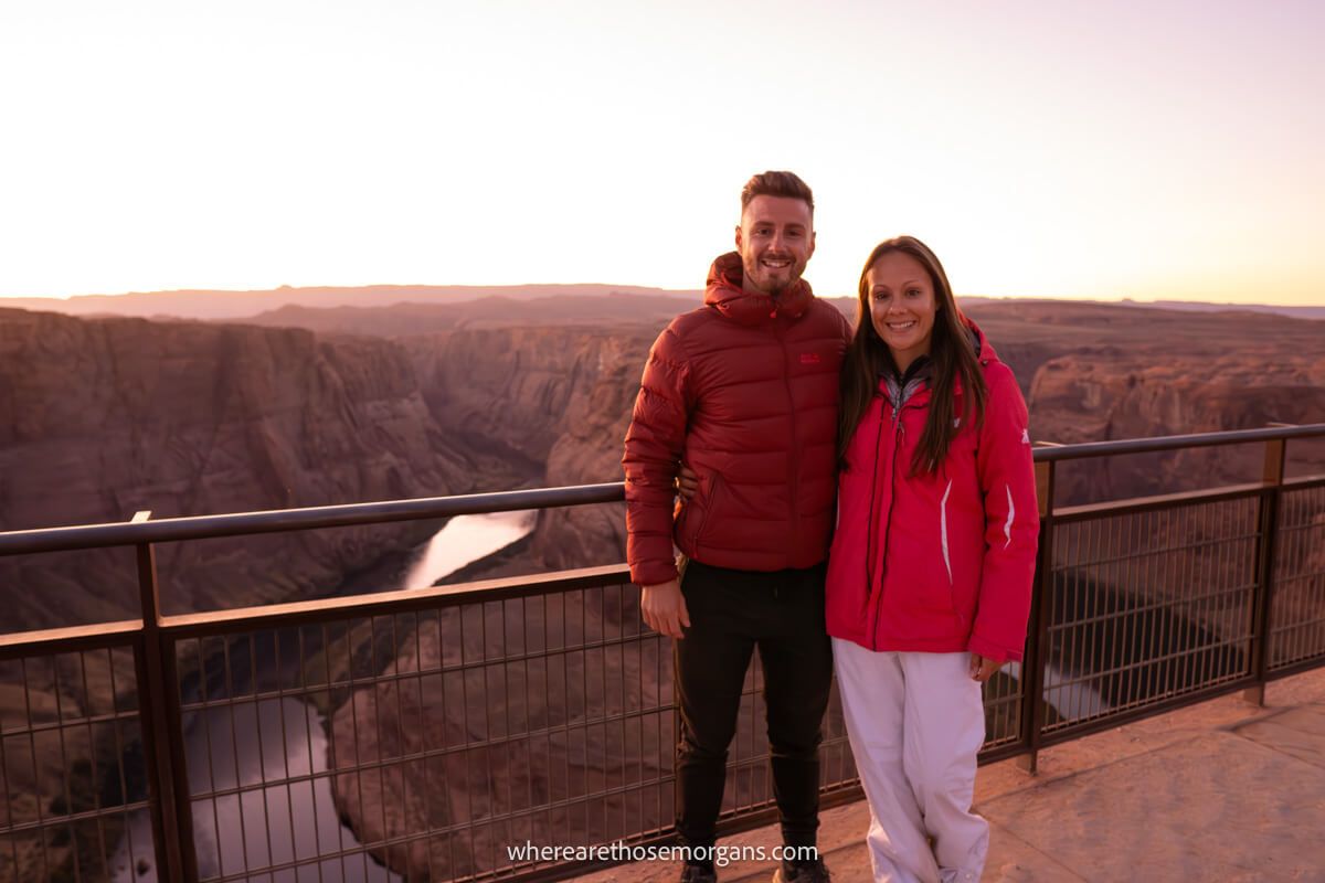 Two tourists in winter clothes standing on a viewing platform next to a protective railing at sunset with views over a U-shaped curve in the Colorado River from above
