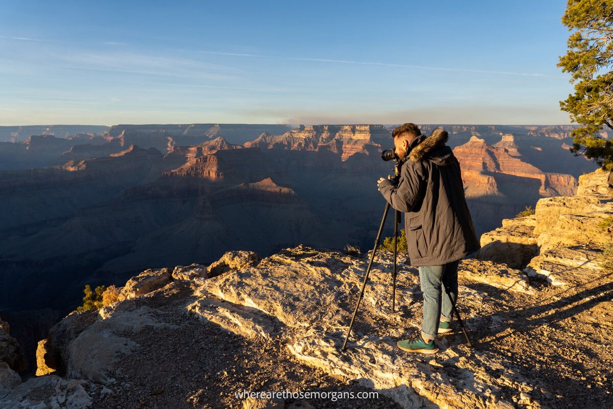 Photographer with camera and tripod in heavy winter coat stood on a rocky outcrop at Hopi Point during sunset