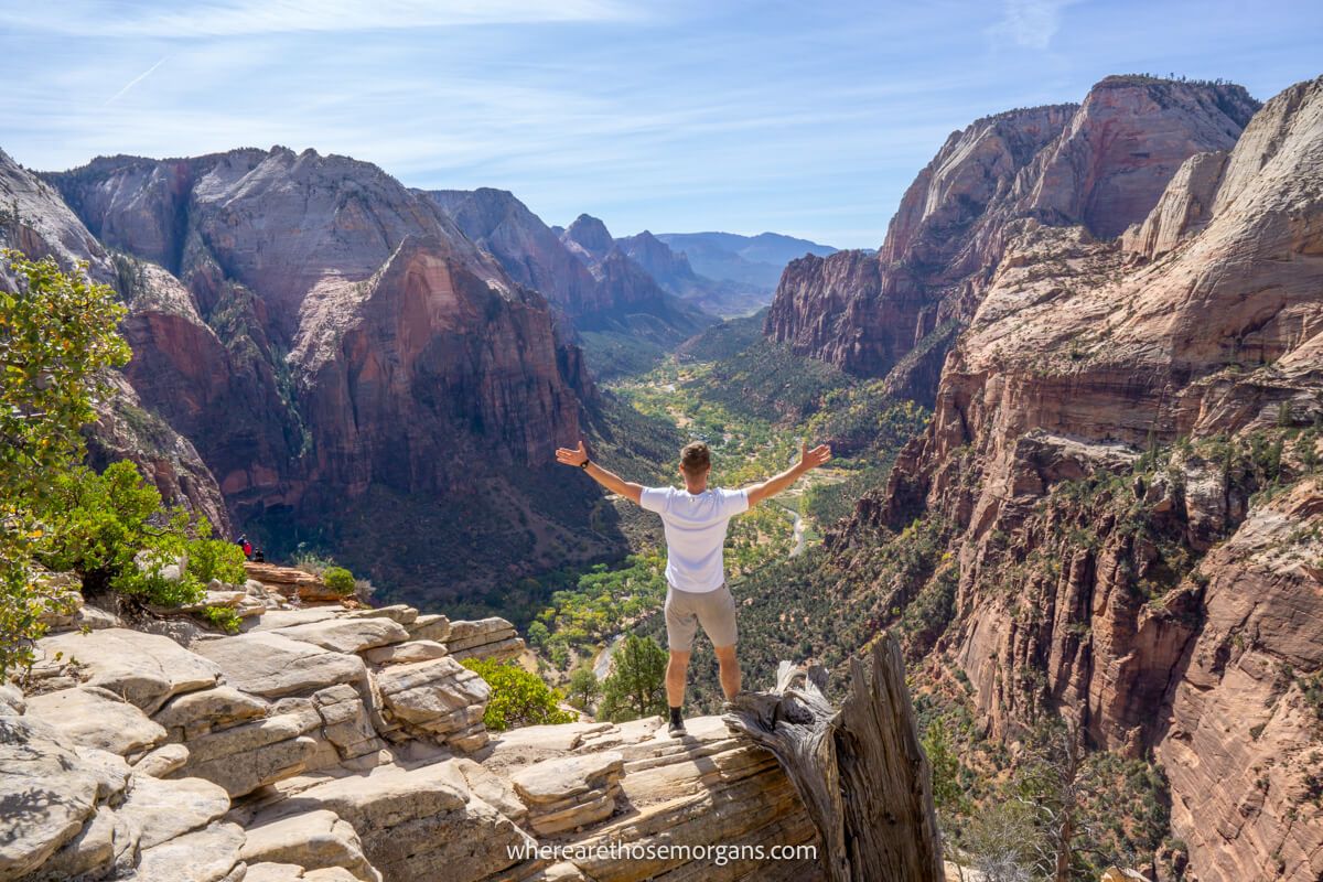 Hiker in white shirt with arms spread wide at the top of a hiking trail called Angels Landing in Zion looking over a valley and canyon walls