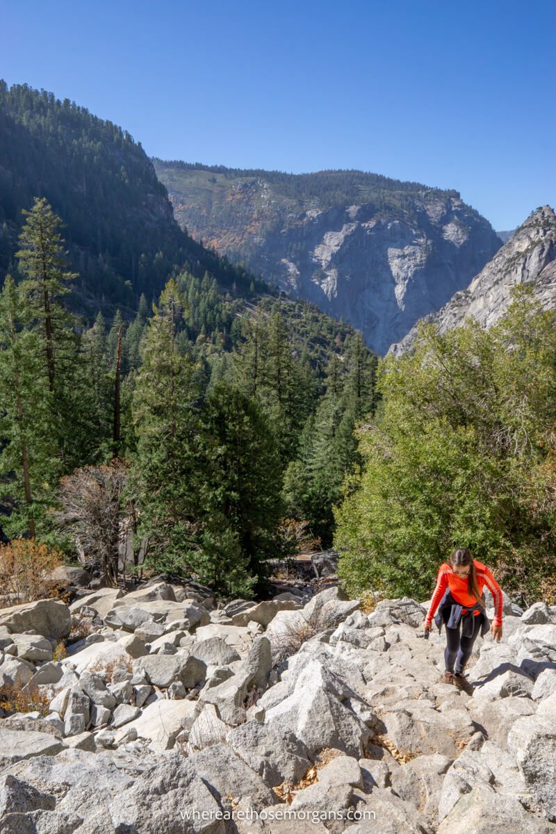 Hiker in red shirt walking up a steep boulder hike with views of trees and granite mountains