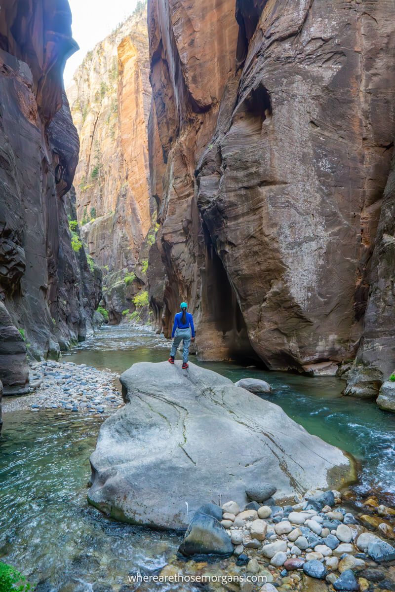 Hiker stood on a huge boulder in the middle of a river flanked by tall slot canyon walls on the Zion Narrows hike