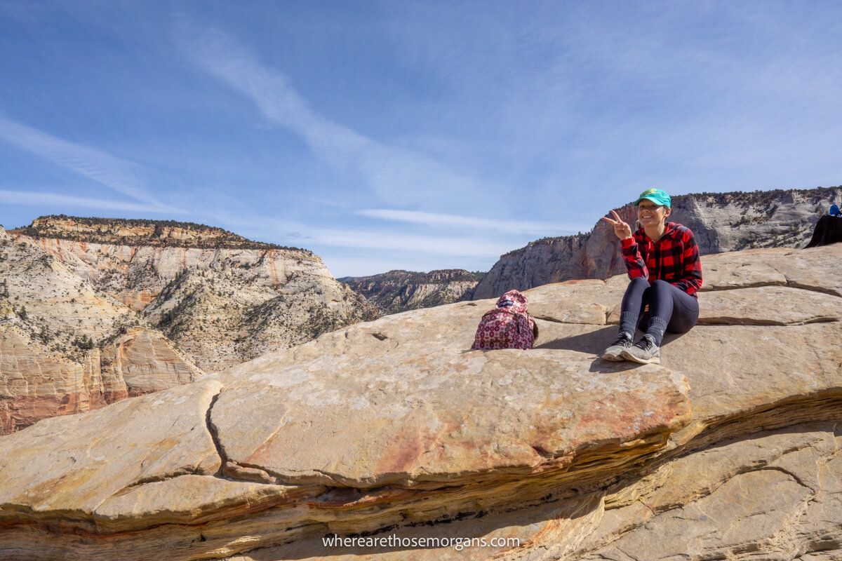 Hiker sat on a rocky summit holding two fingers up in the peace sign at the camera