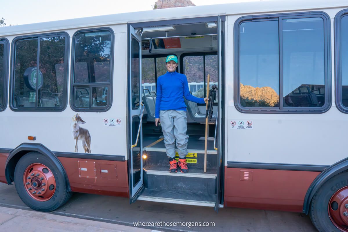 Hiker in blue shirt and green cap holding a wooden pole standing in the doorway of a shuttle bus