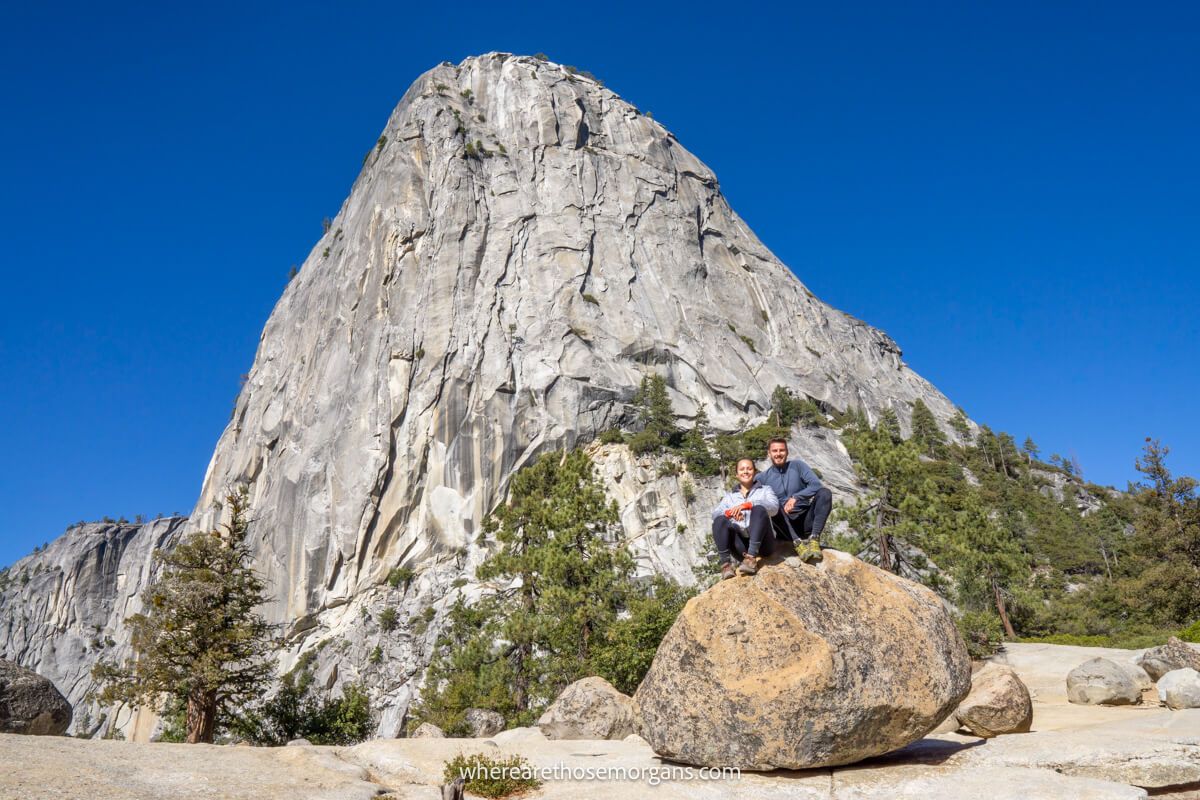 Hiking couple sat together on a boulder in front of Liberty Cap a huge granite monolith on the Mist Trail John Muir Trail loop hike in Yosemite on a clear day with blue sky