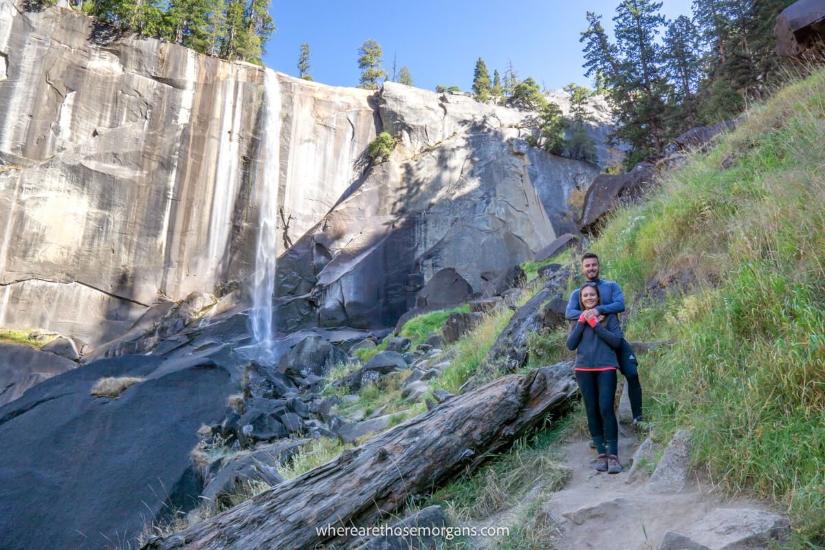 Two hikers embracing on a hiking path next to a log with a wispy waterfall plummeting from a flat rock behind