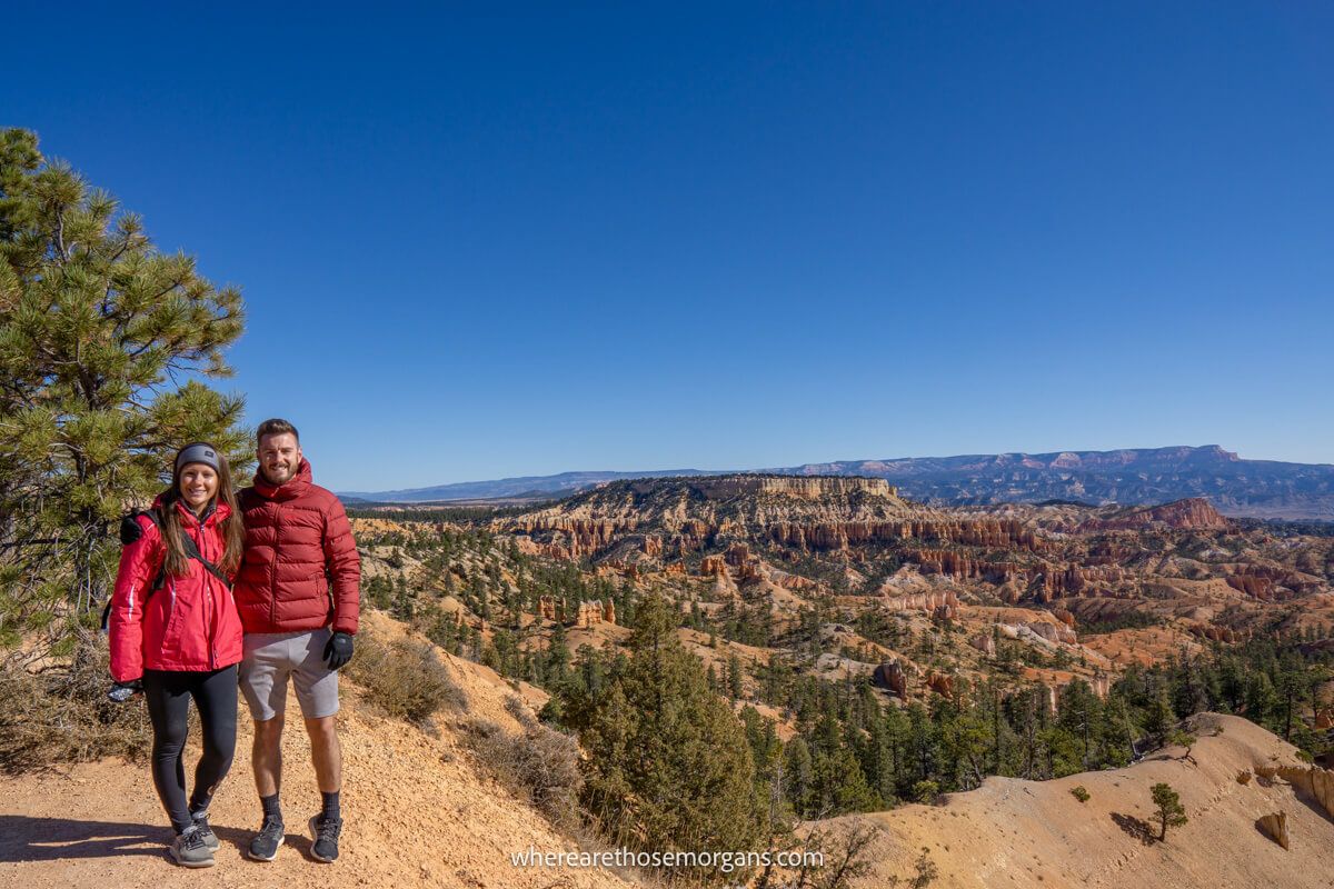 Two hikers stood together in winter coats and gloves on a bright sunny day in Utah for a photo with far reaching views over a sandstone landscape