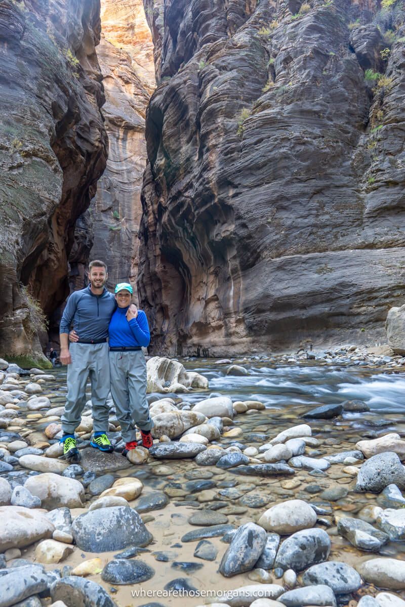 Couple standing together for a photo on small rocks in the middle of a river slot canyon wearing waterproof pants and shoes
