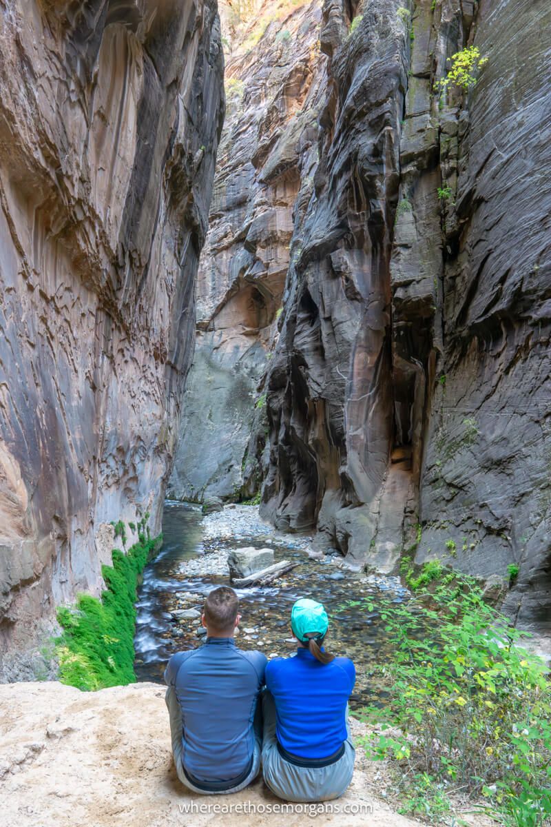 Two hikers sat together looking away from the camera through a slot canyon with a river below