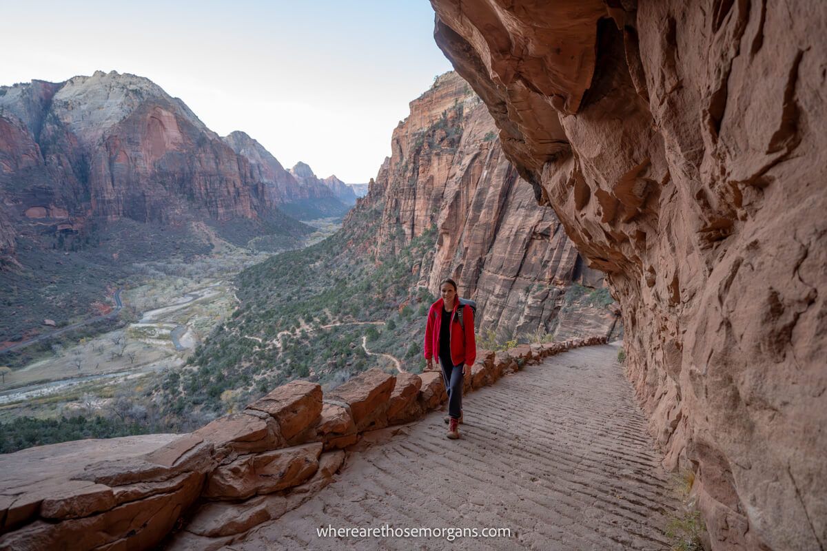 Lone hiker walking up a steep path under an overhanging cliff wall with distant views over a valley flanked by tall canyon walls on the West Rim Trail in Zion