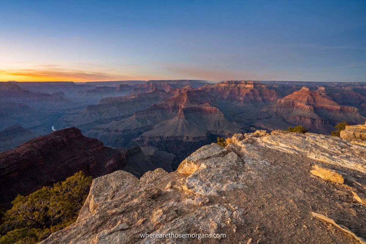 Photo of a sunset over Grand Canyon South Rim with colors in the sky and formations glowing red behind a white rocky outcrop in the foreground