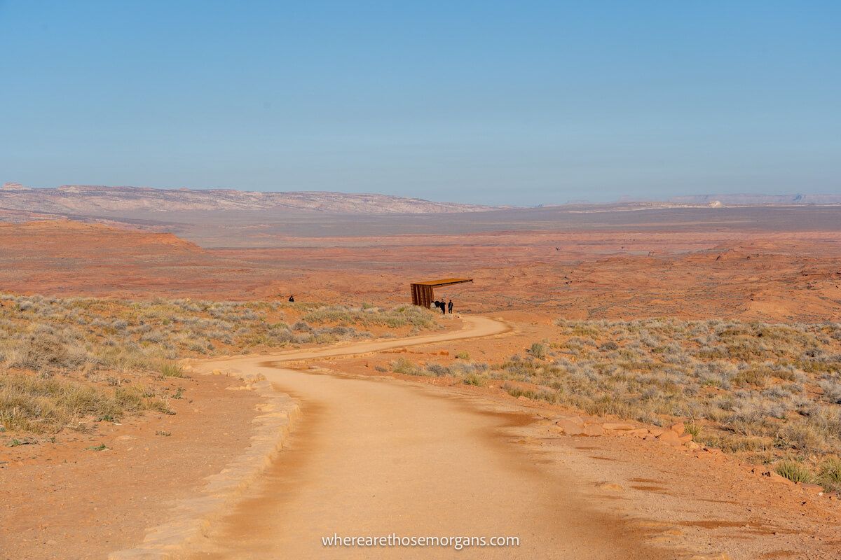 Wide pedestrian trail leading through a sandy desert environment with a small wooden resting area in the distance
