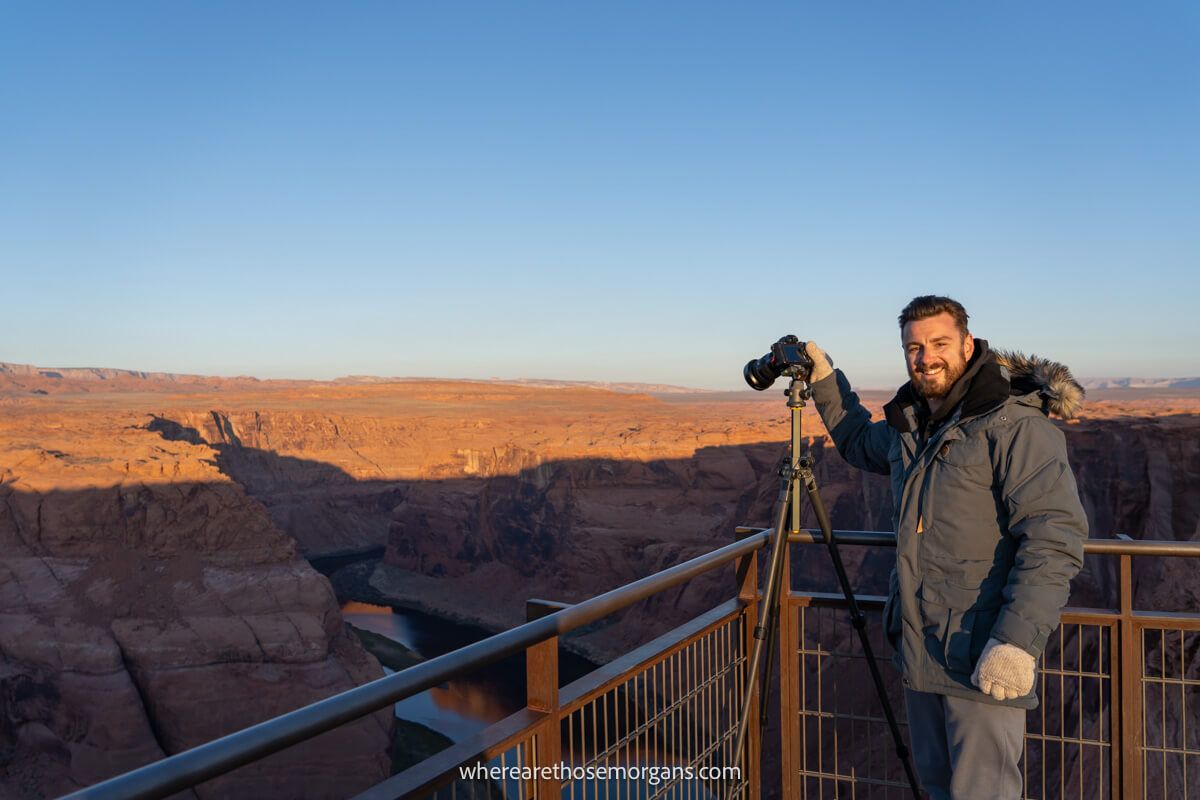 Photographer in winter coat and mittens holding a camera on a tripod next to a fence overlooking a natural rocky landscape at sunrise